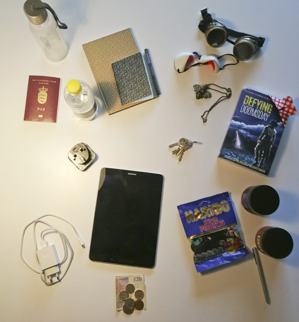 Con Survival Guide: A mess on a table: a waterbottle, a passport, notebooks, jewelry, charger, cash, candy, book, googles 